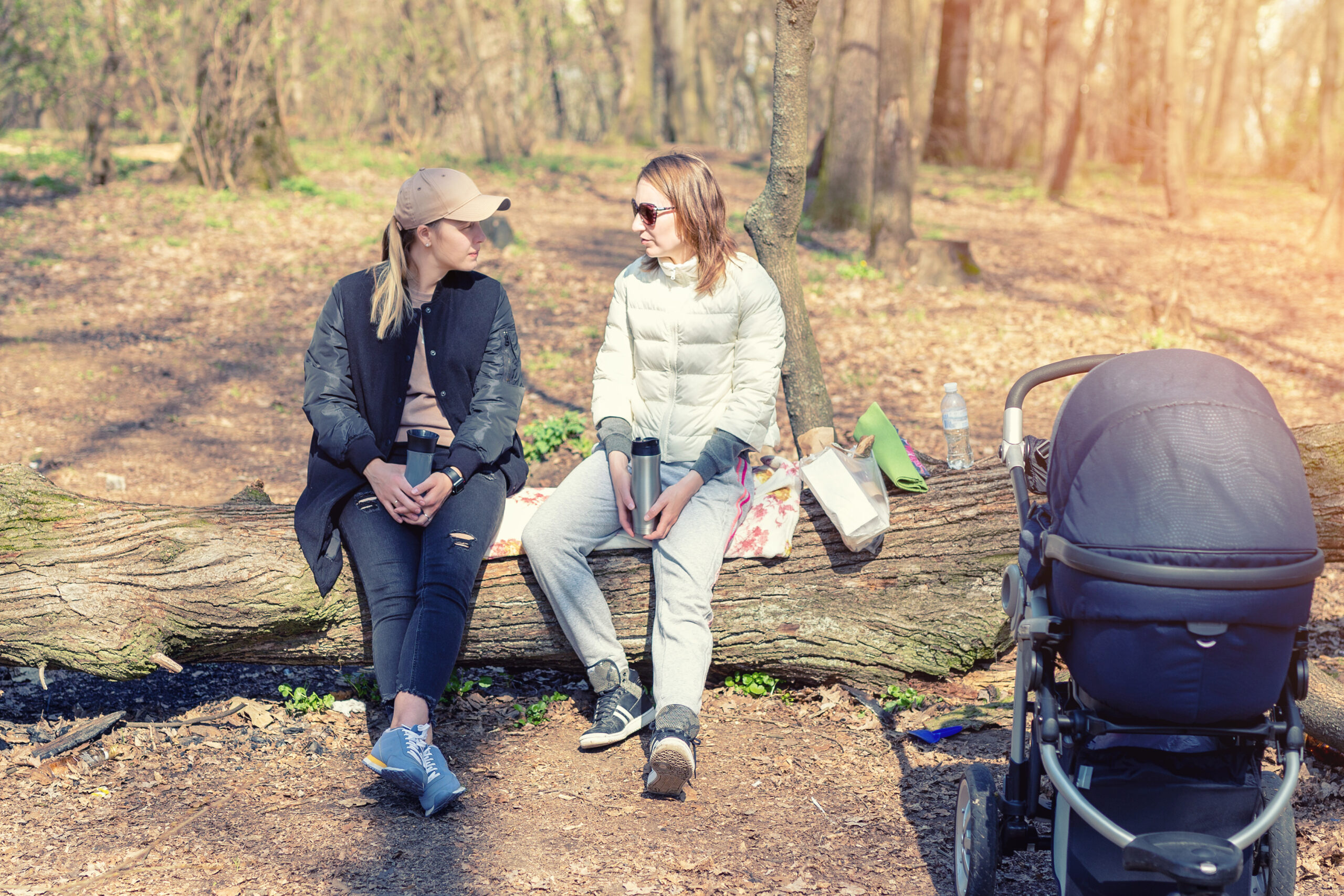 Two women girlfriends sitting on wooden log at forest glade while walking with baby stroller. Young mothers friends talking , communication, gossips and support
